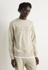 Sudadera de color beige claro con el logo "BOSS" en un tono a juego. Presenta un cuello redondo, puños acanalados y una textura suave. Combinada con pantalones beige.