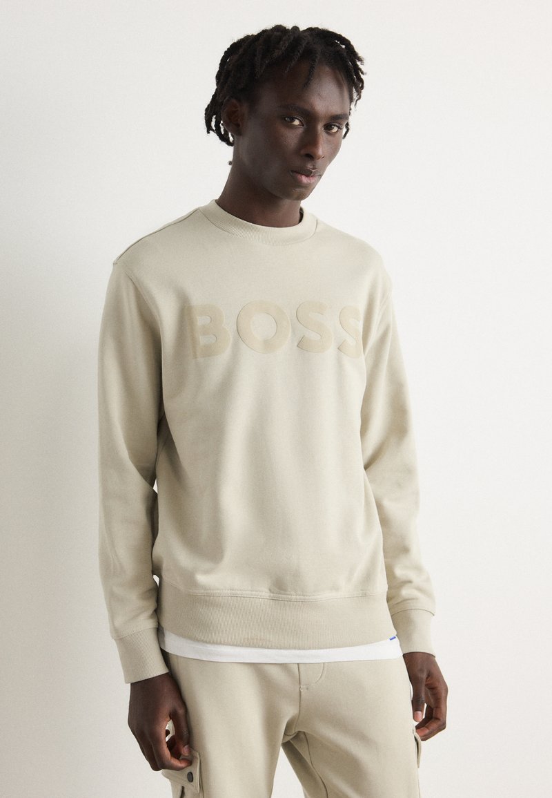 Sudadera de color beige claro con el logo "BOSS" en un tono a juego. Presenta un cuello redondo, puños acanalados y una textura suave. Combinada con pantalones beige.