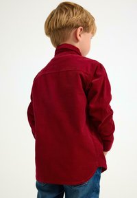 Ung dreng med kort blondt hår iført en langærmet rød cordkjorte og blå jeans, set bagfra mod en ensfarvet lys baggrund.