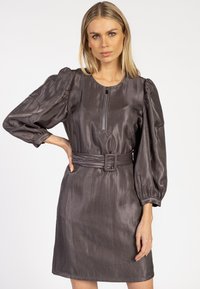 Vrouw met blond haar, die een glanzende donkergrijze jurk met riem draagt, voorzien van aangeknipte lange mouwen en een rits aan de voorkant.