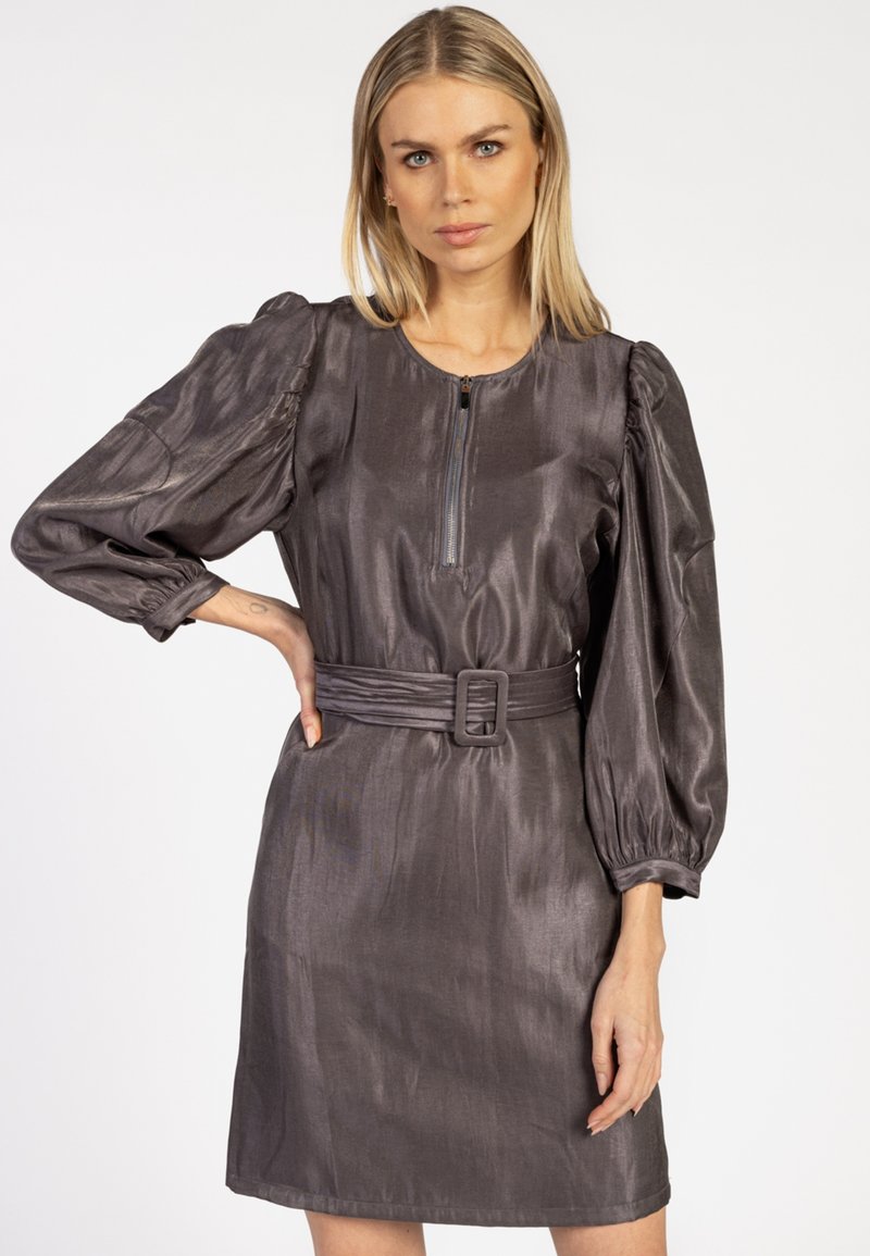 Vrouw met blond haar, die een glanzende donkergrijze jurk met riem draagt, voorzien van aangeknipte lange mouwen en een rits aan de voorkant.