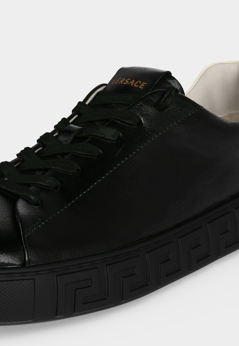 Schwarzer Ledersneaker mit schwarzen Schnürsenkeln, geprägter, gemusterter Sohle und goldenem "Versace"-Logo auf der Zunge, aus einem Winkel gezeigt.