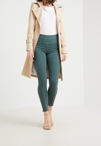 Cappotto trench beige indossato su una maglietta aderente bianca e leggings verdi. I leggings hanno una texture liscia e vita alta, completati da sandali nudi con cinturini.