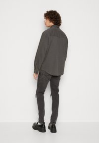 Camisa de manga larga gris oscuro con cuello alzado y puños abotonados, combinada con jeans negros desgastados y zapatos negros gruesos.