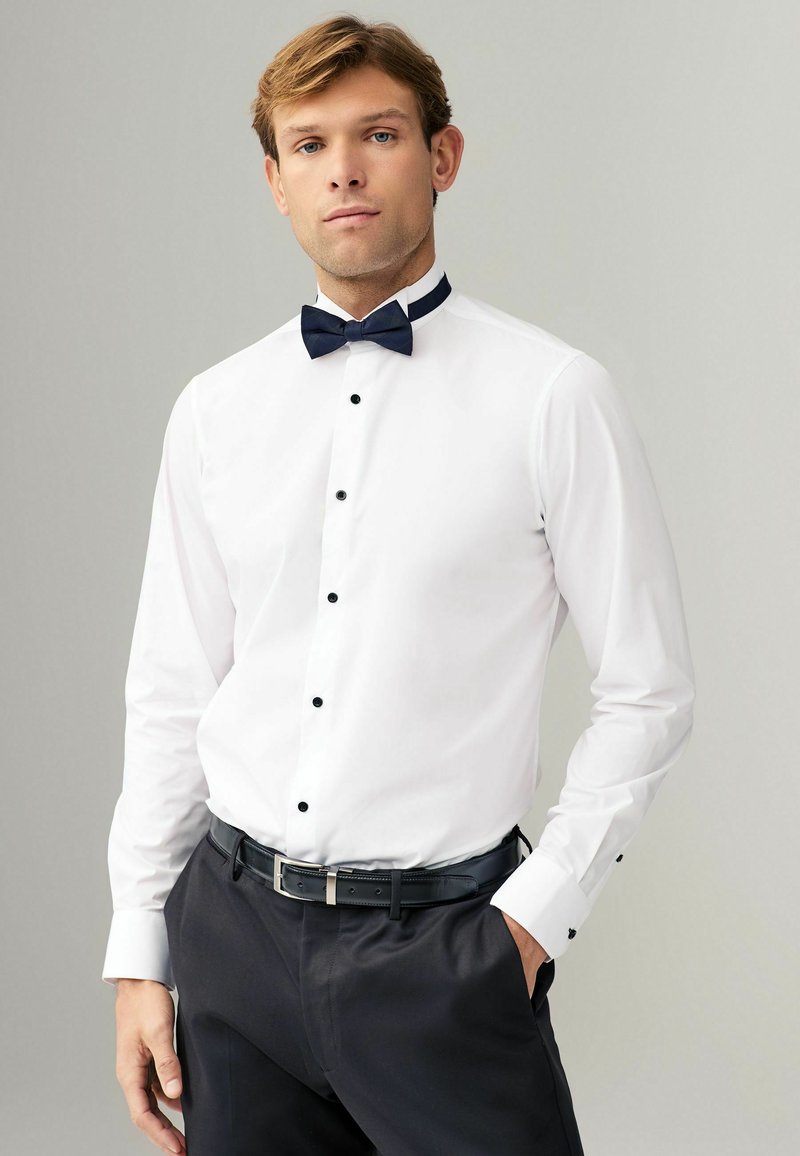 Camisa blanca con botones negros, con una pajarita azul marino y un cuello de contraste; combinada con pantalones oscuros y cinturón negro.