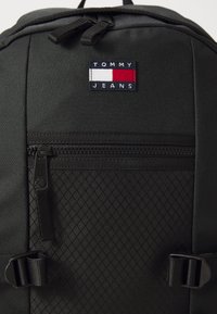Fekete hátizsák tartós anyagból, gyémántmintával, elülső cipzáras zsebbel és Tommy Jeans logóval.
