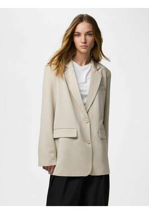 Žena s dugom plavom kosom koja nosi bež sako preko bijele košulje i crne hlače, stoji nasuprot jednobojnoj svijetloj pozadini.