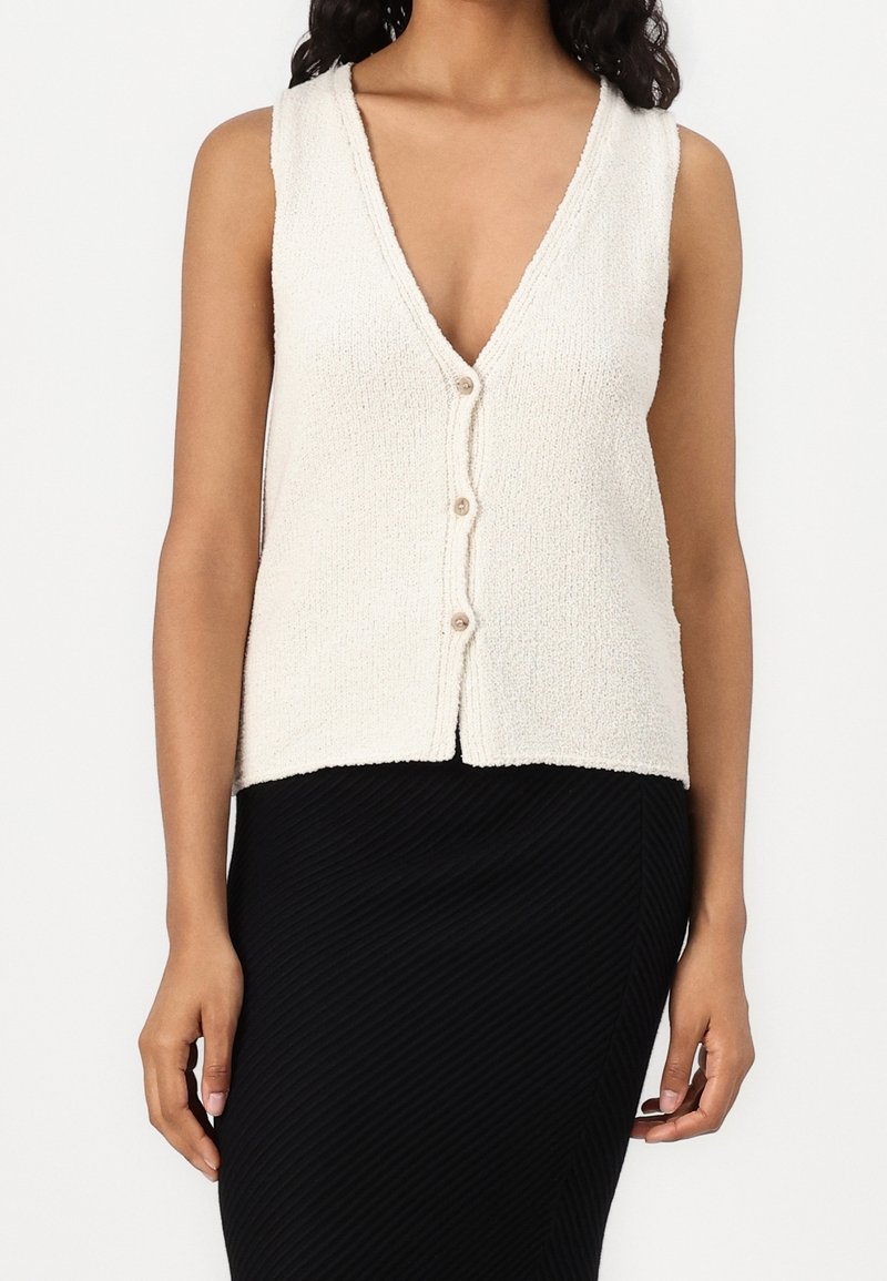 Femme portant un gilet crème texturé sans manches avec boutons et col en V profond, et une jupe noire côtelée sur fond blanc.