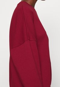 Sudadera burdeos con un corte holgado, mangas largas y una textura suave, con detalles de costura expuesta a lo largo del hombro y los lados.
