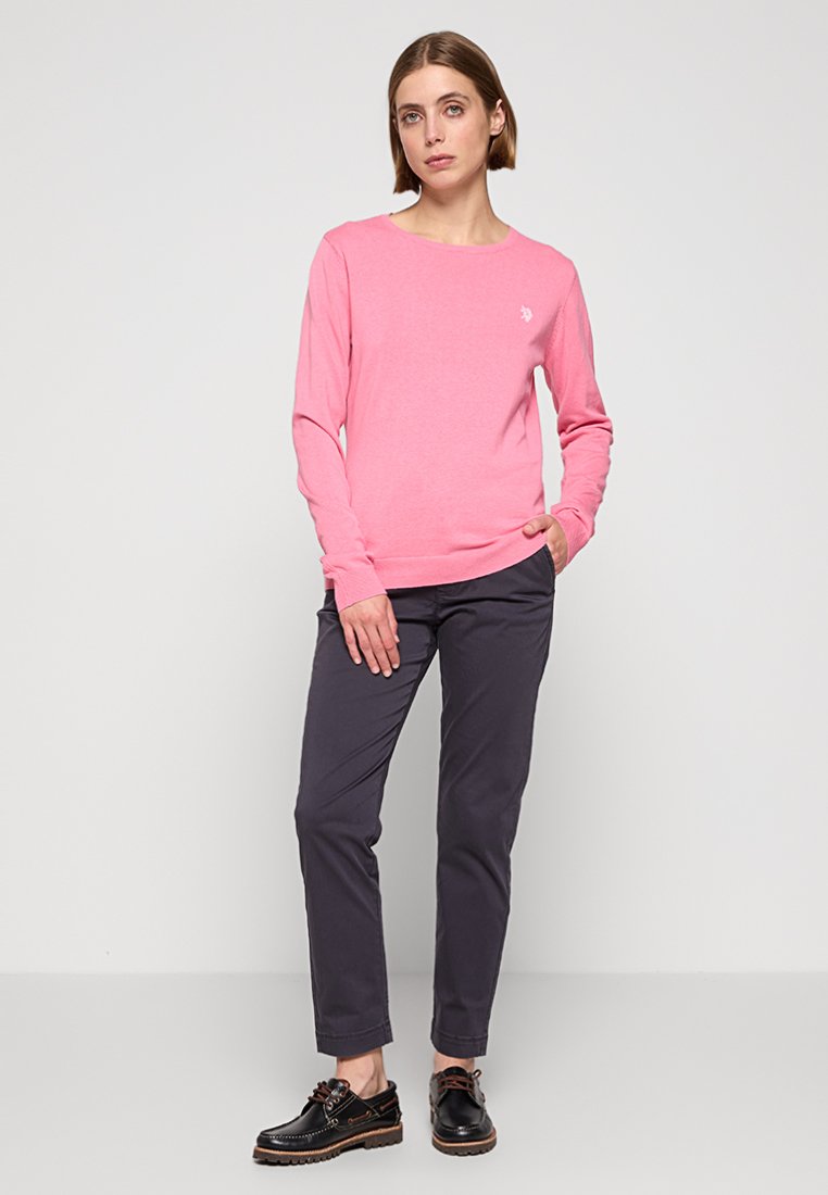 Camicia rosa a maniche lunghe con scollatura rotonda, indossata con pantaloni grigio scuro e scarpe nere, presenta un logo sottile sul petto.