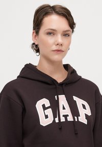 Hanorac maro cu buzunar frontal de tip kangaroo, având un mare logo brodat "G.A.P." în alb și roz. Textură moale a fabricii și glugă cu șnur.
