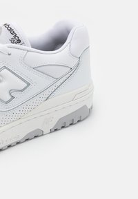 Baskets blanches New Balance 550 montrant le côté et l'arrière avec cuir perforé et détail de semelle en caoutchouc sur fond blanc.