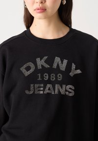 Svart hettegenser med DKNY JEANS-logo utsmykket med sølvfargede rhinestener, med rund hals og en avslappet fasong.