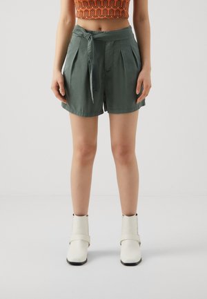 Shorts verts taille haute avec plis, ceinture à nouer et texture lisse. Portés avec des bottines blanches à la cheville.
