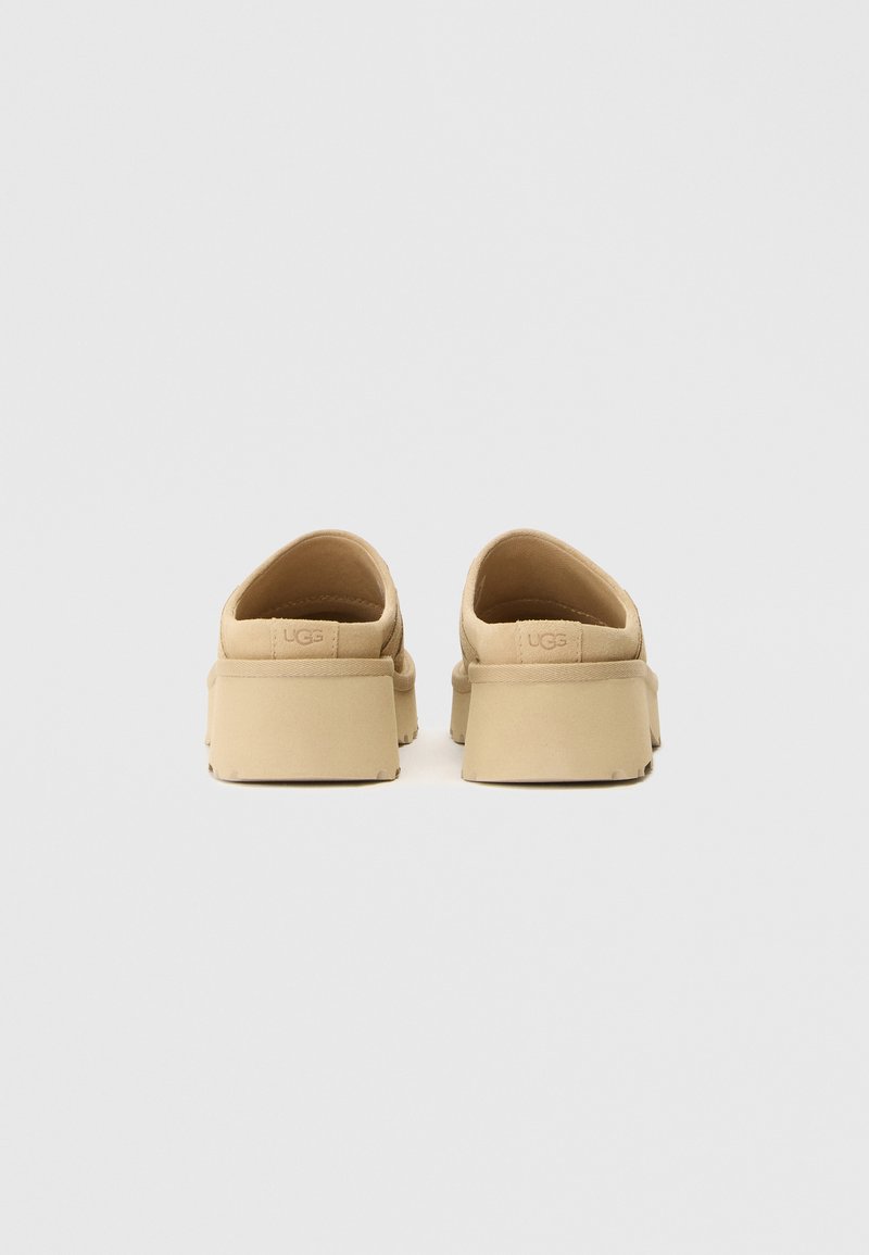 Een paar beige instap schoenen met dikke platformzolen, bekeken vanuit de achterkant op een effen witte achtergrond.