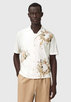 Fehér, rövid ujjú, virágmintás gombos ingben és bézs nadrágban álló férfi, kezét a zsebében tartva, szürke háttér előtt.