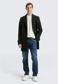 Zwarte wollen jas met grote revers, knoopsluiting, gedragen over een wit shirt met ronde hals en blauwe spijkerbroek; gecombineerd met witte sneakers.