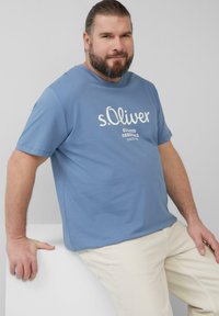 Lichtblauwe katoenen t-shirt met het witte "s.Oliver" logo en tekst. Korte mouwen, ronde hals, gecombineerd met off-white broek, leunend op een witte ondergrond.