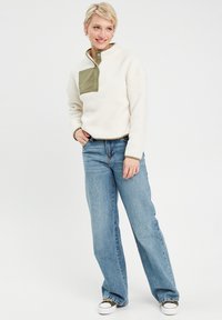 Fleece-Pullover in Creme mit olivgrünem Brustpaneel; verfügt über einen vorderen Druckknopfverschluss, lange Ärmel und eine lockere Passform. Kombiniert mit hellblauen Jeans.
