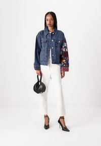 Jeansjacka med blå kropp och flerfärgad patchworkärm, kombinerad med vita vida byxor och svarta klackskor, hållandes en svart väska.