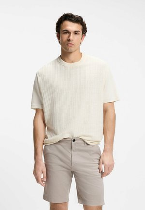 Giovane uomo che indossa un maglione a maniche corte color crema a coste e pantaloncini beige chiaro con trama, in piedi davanti a uno sfondo bianco semplice.