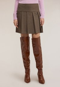 Gonna plissettata a quadri marrone con vita alta, abbinata a stivali marroni lucidi sopra il ginocchio con punta appuntita. La texture del tessuto è morbida.