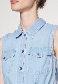 Jasnoniebieska bezrękawnikowa koszula dżinsowa z kołnierzem, dwoma przednimi kieszeniami i srebrnymi zatrzaskami. Gładka tekstura i prosty design.