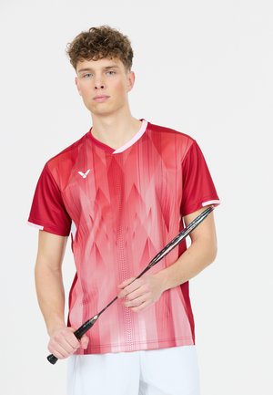 Ung mand i rød sportstrøje holder en badmintonketsjer lodret mod en ensfarvet hvid baggrund.