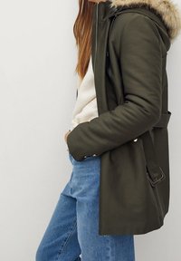 Oliwkowa parka z kapturem wykończonym futrem, wodoodporny materiał i regulowany pasek. Połączona z jasnoniebieskimi dżinsami i beżowym swetrem.