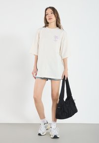 Overdimensjonert kremfarget t-skjorte med lilla palmegrafikk, kombinert med denimshorts og svart skulderveske. Modellen har på seg hvite og sølvfargede sneakers.