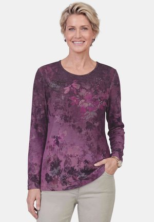 Vrouw die glimlacht, draagt een paarse bloemen top met lange mouwen met kleine strass-steentjes, beige broek en een polshorloge, staand tegen een effen achtergrond.