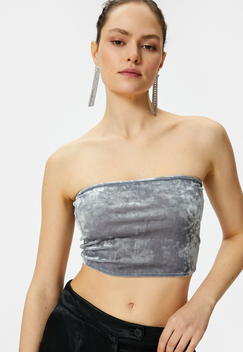 Top corto sin mangas de terciopelo gris con una textura recogida. La modelo lleva pendientes largos y brillantes de plata. Pantalones de satén negros de tiro alto visibles por debajo.