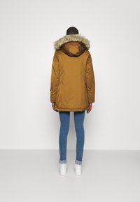 Persoon in een bruine parka met een bontafgepelde capuchon, blauwe spijkerbroek en witte sneakers, staand met de rug naar de camera tegen een effen achtergrond.