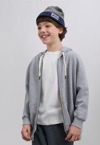 Grijze hoodie met ritssluiting en crèmekleurige trekkoorden, gedragen over een wit shirt. Grijze gebreide muts met blauwe rand en logo. Getextureerde grijze broek.