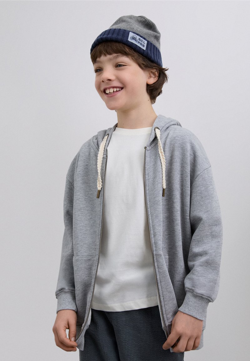 Grijze hoodie met ritssluiting en crèmekleurige trekkoorden, gedragen over een wit shirt. Grijze gebreide muts met blauwe rand en logo. Getextureerde grijze broek.