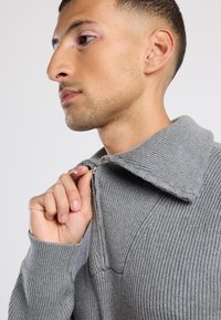 Hombre con cabello corto y una ligera barba de tres días subiendo la cremallera de un suéter de punto acanalado gris con un cuello ancho.