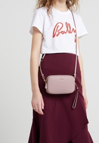 Olkalaukku pehmeää vaaleanpunaista nahkaa, jossa on sileä pinta, kultaiset hardwaret ja suorakulmainen muoto, yhdistettynä burgundinpunaisiin hameeseen ja valkoiseen T-paitaan.