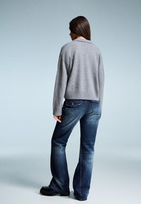 Grauer gestrickter Pullover mit geripptem Saum, kombiniert mit blauen Schlagjeans. Die Jeans haben eine ausgebleichte Optik und sichtbare Nähte.