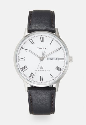 Reloj Timex Waterbury con esfera blanca, números romanos, manecillas plateadas y correa de piel negra con costuras en contraste.