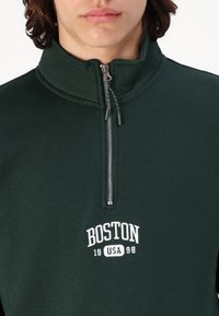 Mørkegrønn hettegenser med høy krage og glidelås, prydet med hvit brodering som leser "BOSTON 19 USA 98" over glidelåsen. Behagelig tekstur.