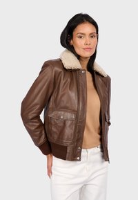 Chaqueta bomber de cuero marrón con cuello de borrego crema, bolsillos frontales y puños acanalados. Textura suave, cierre frontal con cremallera y diseño ajustado.