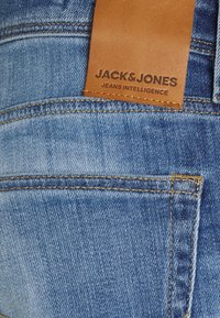 Siniset farkut, joissa on vaaleanruskea nahkainen merkki, johon on painettu "JACK & JONES" ja "JEANS INTELLIGENCE". Näkyvissä ompelut ja taskun yksityiskohdat.