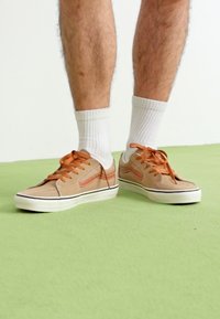 Beige velúrbőr sportcipők narancssárga részletekkel és fehér gumitalppal. Narancssárga cipőfűzőkkel megkötve. Fehér, rövid szárú zoknival viselve. Zöld padló.