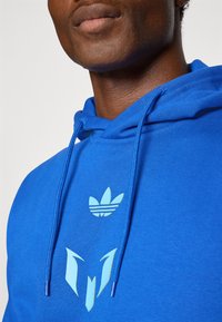 Sweatshirt azul com capuz feito de tecido macio, apresentando um logótipo gráfico ciano no peito e capuz, com cordões combinando e detalhes texturizados.