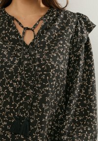 Blusa nera con stampa floreale, scollatura a chiave con lacci e rouches sulle spalle. Realizzata in tessuto morbido con un accento a frangia al centro.