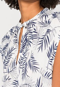 Korte mouwen blouse van witte stof met marineblauwe bladranden. Beschikt over een keyhole-halslijn met een strikdetail en een licht gestructureerd oppervlak.