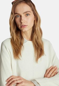 Biały sweter z prążkowanej tkaniny z pasiastym kołnierzem, noszony przez osobę z długimi, falującymi włosami, która ma na rękach warstwowe złote pierścionki.