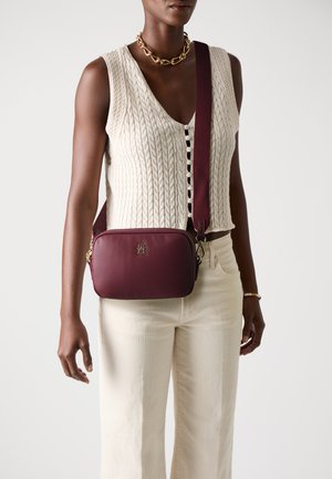 Bordeauxfarbene Leder-Crossbody-Tasche mit goldenen Beschlägen, rechteckiger Form und strukturiertem Schultergurt, vor einem hellen Hintergrund getragen.