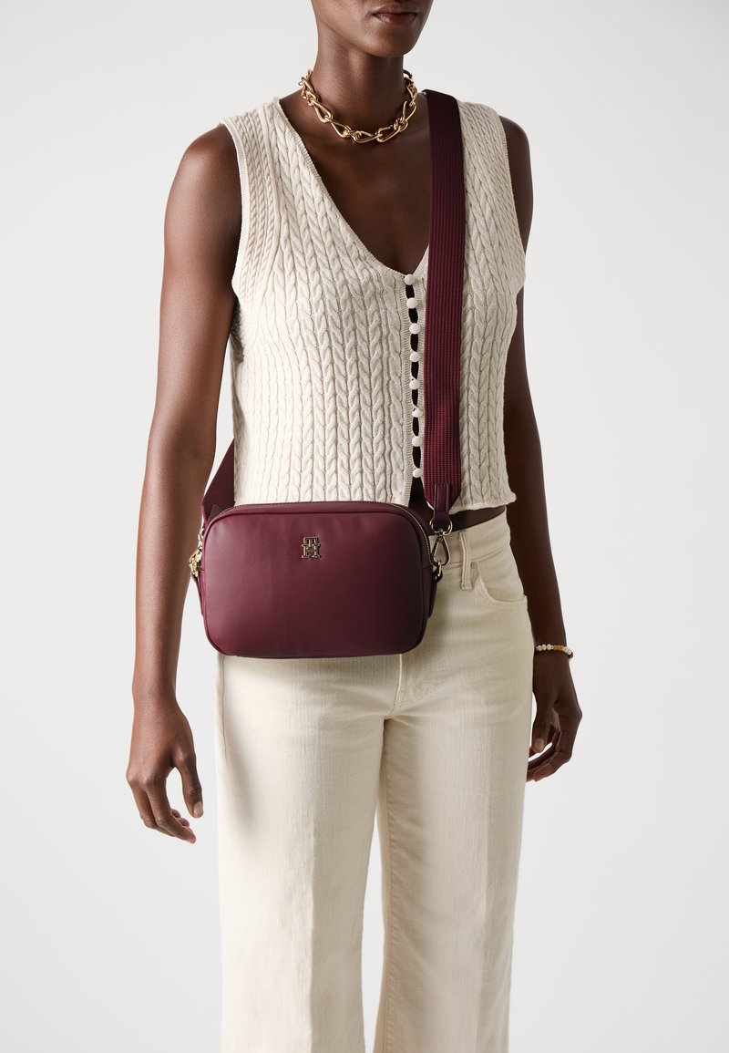 Bordeaux leren crossbody tas met gouden hardware, rechthoekige vorm en een getextureerde schouderriem, gedragen tegen een lichte achtergrond.