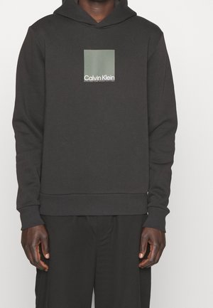 Mand iført sort Calvin Klein hættetrøje med en grøn firkant og hvidt logo på brystet, kombineret med sorte bukser.